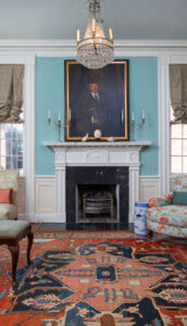 Fireplace in a historic house with detailed molding, antique chandelier, and family heirloom painting above the mantle.