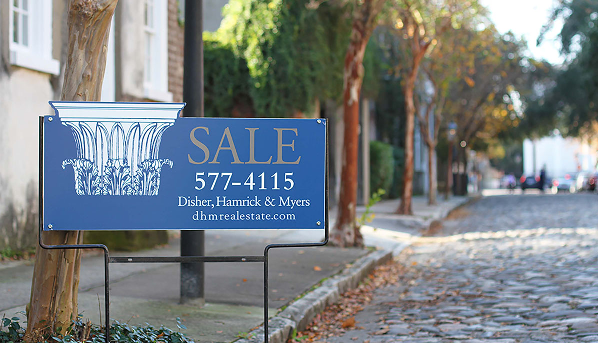 View of cobblestone street in historic downtown Charleston with home for sale sign by Disher, Hamrick & Myers